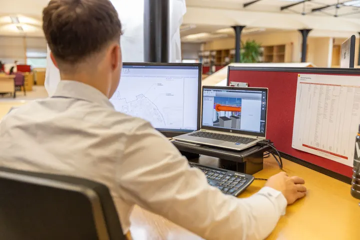 Engineer at two screens showing engineering drawings, seen from behind